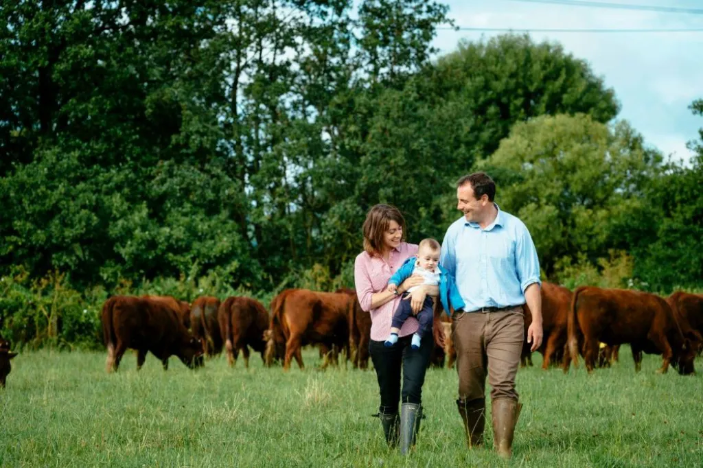 Farmer and wife, Neil and Sally Griggs, holding young son in field with herd of cows in background